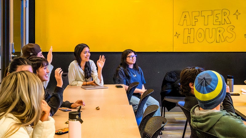 students raise their hands in a classroom in front of a large yellow wall where someone has written "After Hours"