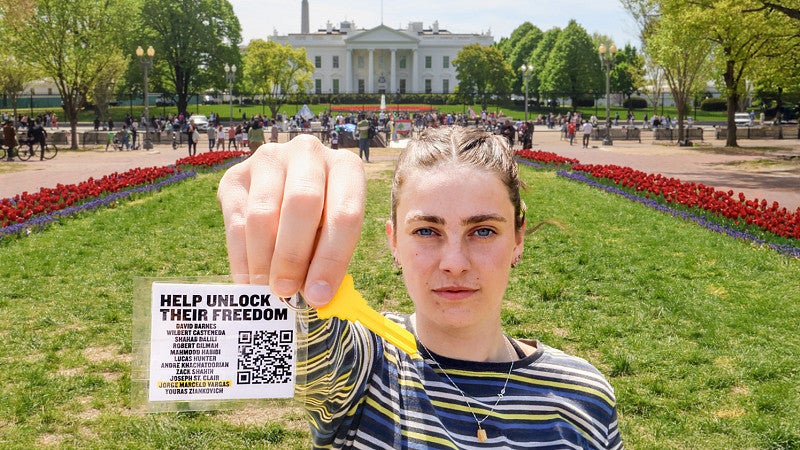 Maren Fullerton holds a white tag attached to a bright yellow key close to the camera; a green lawn and the White House are in the background
