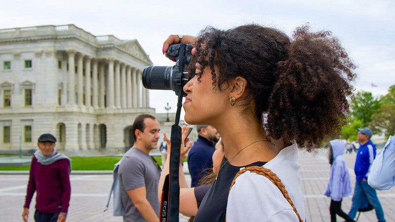 A student with curly dark hair holds a camera up to her eye