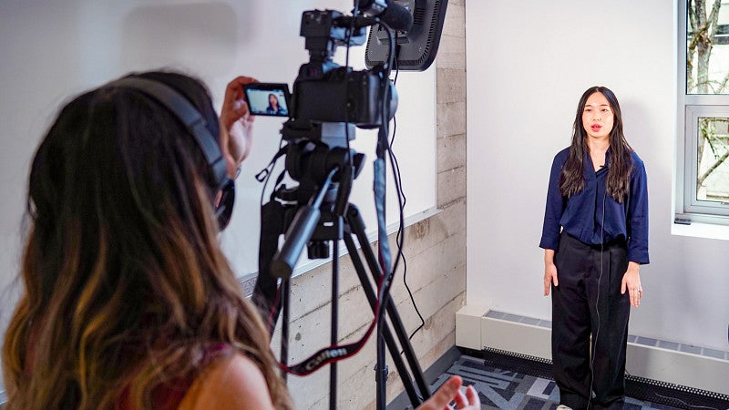 Cing Dim stands behind a video camera filming Mary Pham in a classroom setting