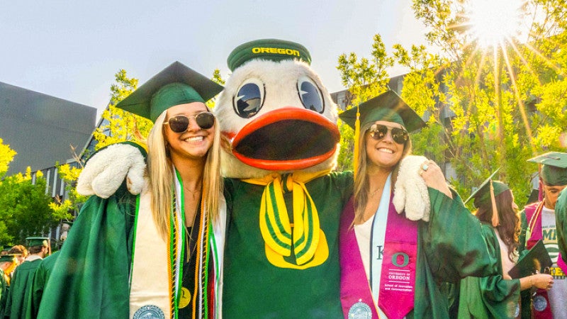 two students in graduation caps and gowns pose with the University of Oregon duck