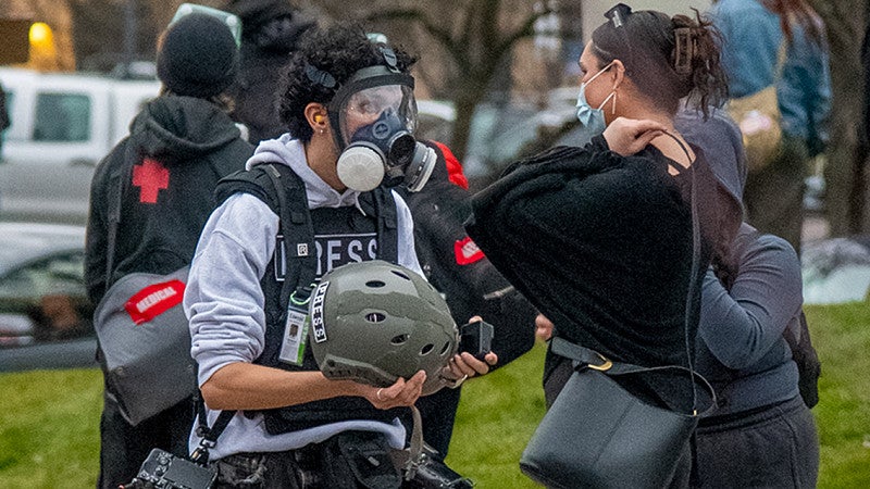 a student photographer wearing a gas mask and carrying a helmet at a protest