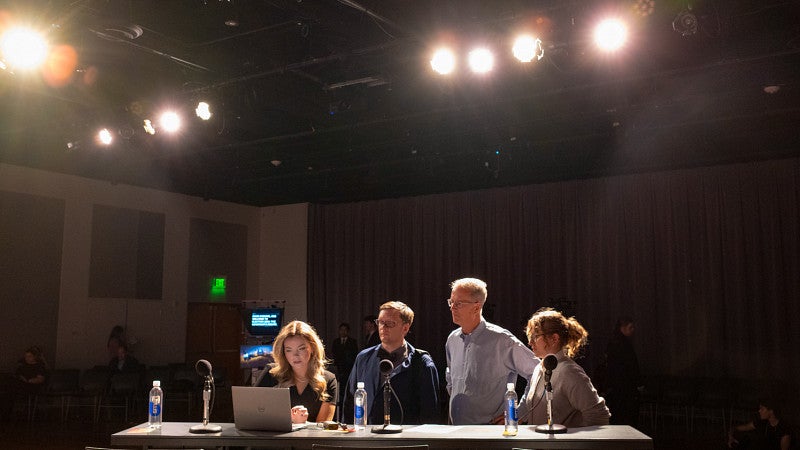 the moderator and panelists for an oregon gubernatorial debate prepare in a dark room lit with studio lights