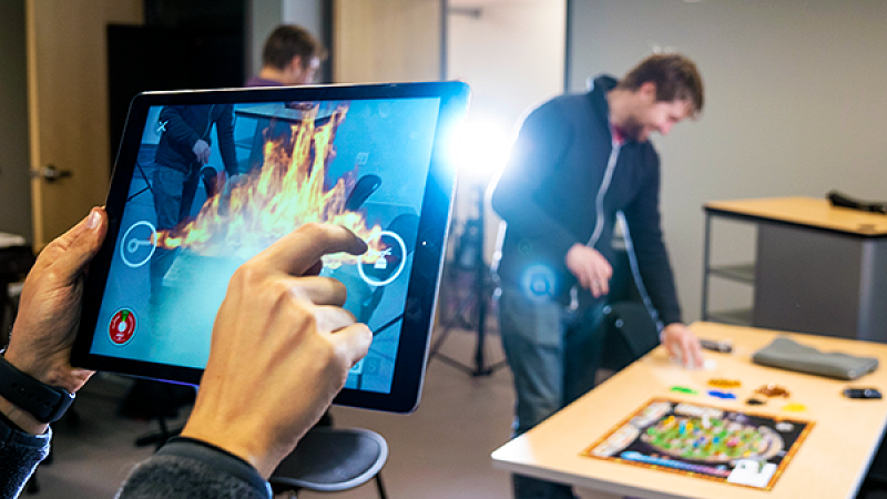 a person clicks on immersive elements on an ipad while another person in the background sets up an immersive game board