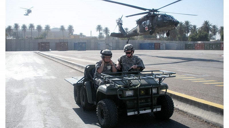 Two people drive a military vehicle while a helicopter hovers overhead.