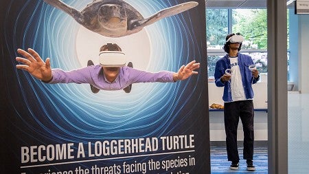a student wearing VR gear stands behind a poster that says "Become a Loggerhead Turtle; Experience the threats facing the species in this interactive virtual reality simulation"