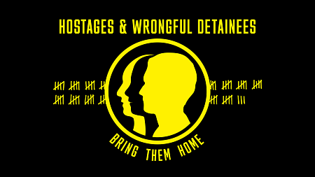 the official US Hostage and Wrongful Detainee Flag which shows a yellow design on a black background. The design includes faces in silhouette, enclosed in a circle, in front of tally marks with the text "Hostages and Wrongful Detainees" at the top and "Bring Them Home" at the bottom 