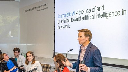 Seth Lewis presents at a microphone from a panel of experts. A screen behind him says "Journalistic AI = the use of and orientation toward artificial intelligence in newswork."