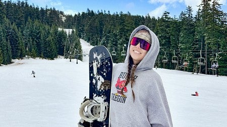 Sydney Seymour wears ski goggles and holds a snowboard while standing in front of a ski lift rising amid evergreen trees