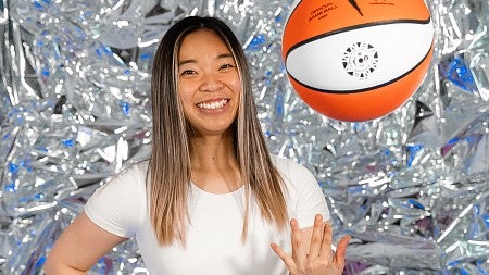 Carly Ebisuya poses for a portrait in front of a shiny silver background while tossing a basketball in the air 