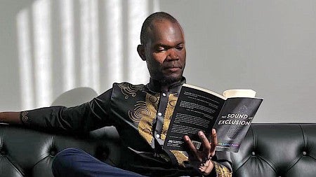 Robert Apiyo reads a book while sitting on a couch with filtered light across his face