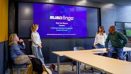 four people flank a large digital screen showing a presentation about the MusoLingo app
