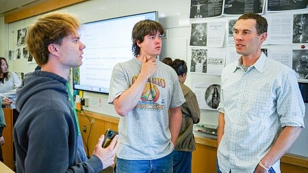 three people stand in a classroom in front of a whiteboard covered in printouts of magazine spreads