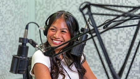 a student looks off camera and smiles while sitting in a podcast recording studio and wearing headphones