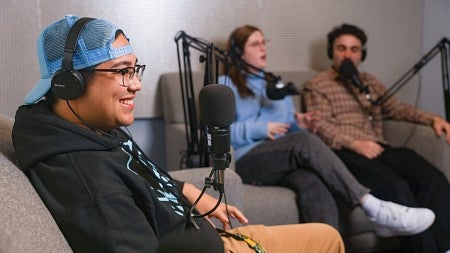 three students sit on couches in the University of Oregon School of Journalism and Communication's podcasting studio with microphones in front of them