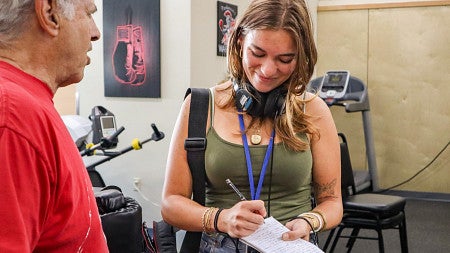 Julia Boboc has headphones around her neck and looks down and smiles as she takes notes while interviewing a person in a red shirt 