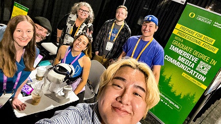 group selfie shot of seven people at a conference display booth with a green and yellow banner advertising the University of Oregon's Graduate Degree in Immersive Media Communication