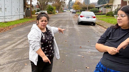 a woman stands on a street pointing towards a parked car while another woman looks on