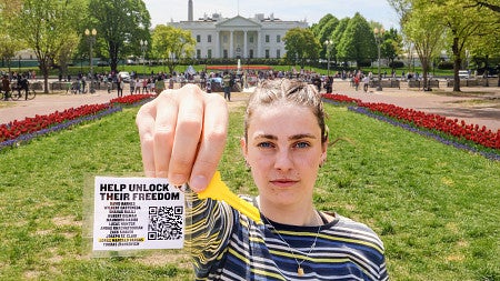 Maren Fullerton holds a white tag attached to a bright yellow key close to the camera; a green lawn and the White House are in the background