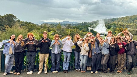 several students make their hands into the shape of an O while posing in New Zealand with a lush green forest landscape them