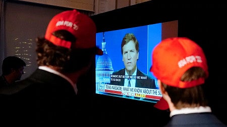 two people wearing red MAGA hats watch Tucker Carlson on a TV screen