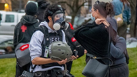 a student photographer wearing a gas mask and carrying a helmet at a protest