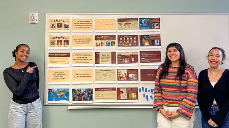 three students pose next to printed slides posted on a whiteboard