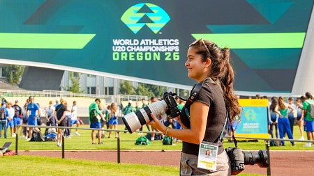 A young woman holds a camera at a track event with the logo of the World Athletics U20 Championships in the background