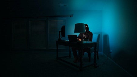 A masked figure sitting in a dark room, illuminated by a blue screen.