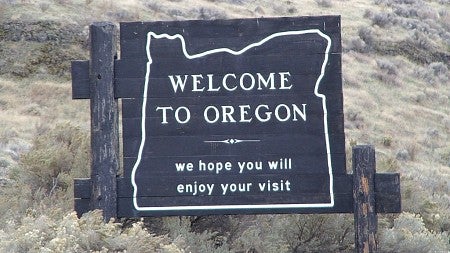 A wooden 'Welcome to Oregon' sign stands in a field, wishing that travelers enjoy their visit (by rustejunk). 