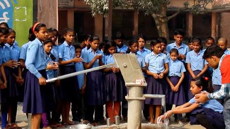 Children in uniform stand around a water pump while one student uses the running water to wash their hands.