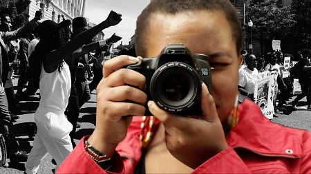 Photographer holds camera in front of graphic with people protesting.