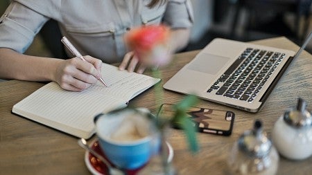 Person wearing a beige button down shirt writing in their notebook while sitting in front of a laptop.