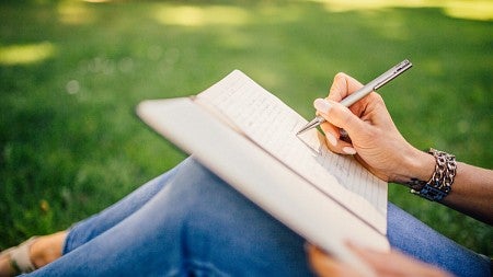 Person wearing blue jeans sitting in the grass writing a story in their notebook.