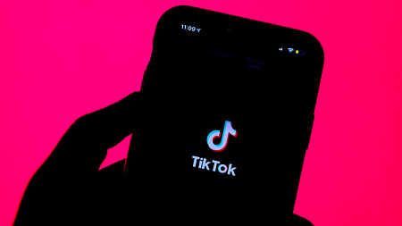 A hand holds a phone with the tiktok logo on screen in front of a pink background.