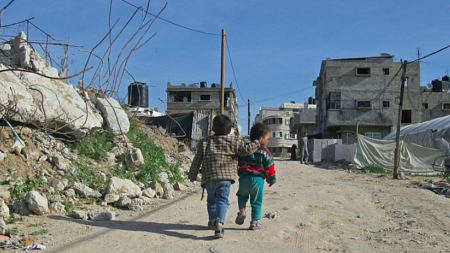 Two children walking away from camera on dirt road towards a small cluster of buildings