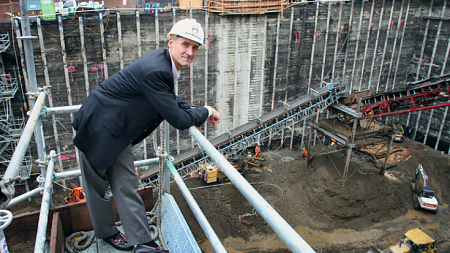 Bart Eberwein, wearing a black and grey suit, stands on scaffolding in front of a construction site - Photo by Cathy Cheney
