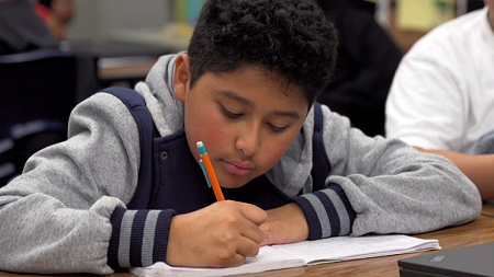 A student wearing a navy and grey sweatshirt writes in a notebook.