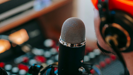 A person wearing an orange beanie speaks into a microphone as they sit in front of a sound mixer (By Anna Pou from Pexels).