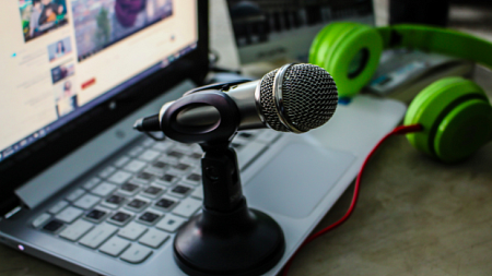 A microphone sits on top of an illuminated laptop next to a set of green headphones (By samer daboul from Pexels).