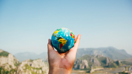 A person holds a small, colorful globe in their outstretched hand in front of a mountainous landscape (By Porapak Apichodilok from Pexels). 