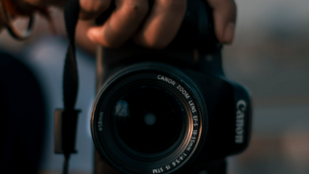 A person wearing a blue and orange shirt holds a camera out in front of them ( By Lensmagicians from Pexels).