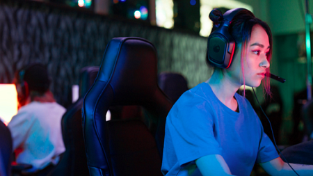 A female gamer wearing a blue shirt looks at a gaming monitor, her face illuminated by the screen (By Ron Lach from Pexels).