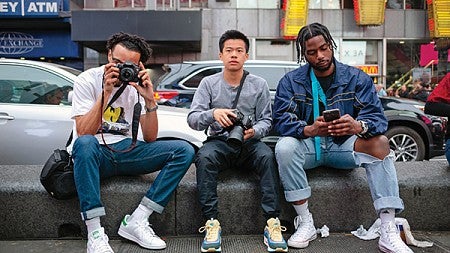 Three male students sitting on a curb taking photos in New York City