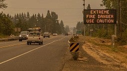 Cars drive along a highway during extremely smoky conditions; a sign next to the road reads "extreme fire danger, use caution"
