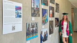 Ally Thomas, wearing her graduation cap and gown, stands next to her photo exhibit in Allen hall