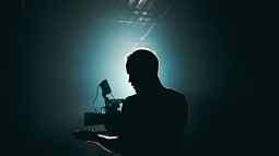 A videographer holding a video camera is backlit in a dark room (By Kyle Loftus from Pexels).