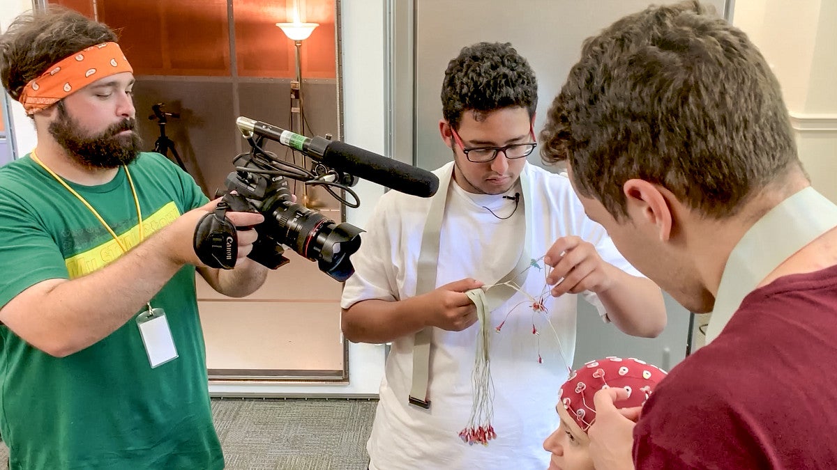 a person wearing a green shirt uses a camera to film three other people conducting a research experiment
