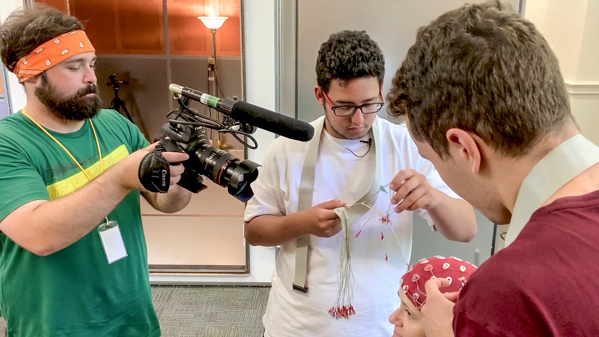  a person wearing a green shirt uses a camera to film three other people conducting a research experiment