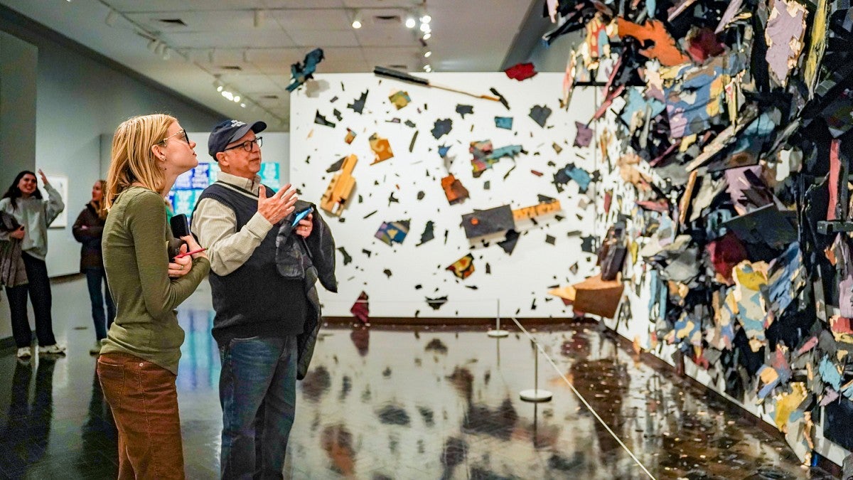 Dave Koranda and a student consider a large scale art piece full of colorful shapes and fragments that looks as if it's exploding across the walls